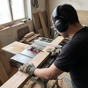 Joiner wearing black electronic Bluetooth ear defenders and PPE gloves while using a table saw in a workshop