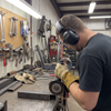 Fabricator wearing black electronic Bluetooth ear defenders and PPE gloves while cutting steel with an angle grinder