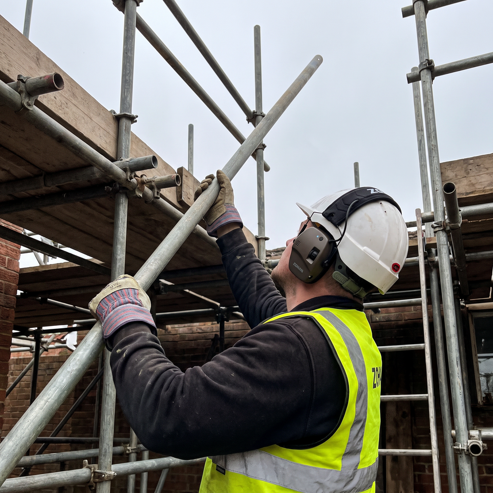 Scaffolder wearing NRR22 electronic Bluetooth ear muffs and PPE gloves while lifting a scaffold tube