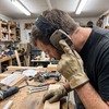 Tradesman pressing the ear cup button on NRR22 electronic Bluetooth ear muffs, wearing PPE gloves in a workshop